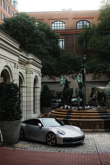 A silver Porsche 911 parked beside a bronze horse fountain in a brick courtyard