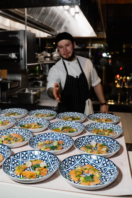 A chef reaching across rows of blue and white plates of salmon crudo in an open kitchen