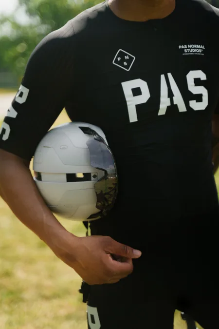 A cyclist in a black Pas Normal Studios kit holding a helmet at their hip