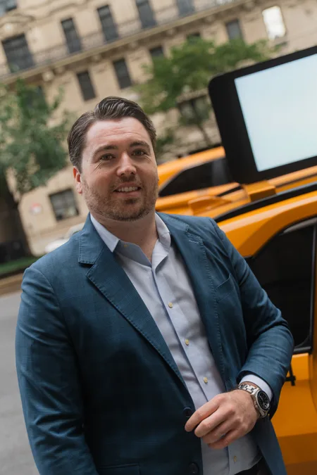 Ned in a blue blazer smiling beside a New York City yellow taxi