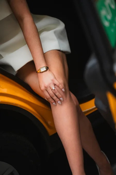 Close-up of Alison's engagement ring and gold watch on a hand resting on a knee against a New York City taxi