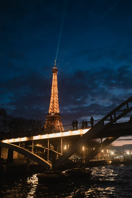 The Eiffel Tower lit at dusk beyond a footbridge over the Seine